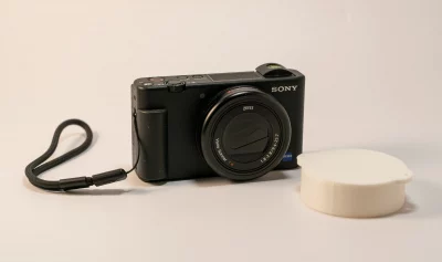 Sony ZV-1 Kompaktkamera mit danebenliegender 3D-gedruckter weißer Schutzkappe, entworfen in Tinkercad. Das 3D Modell steht auf Thingiverse zum Download bereit.