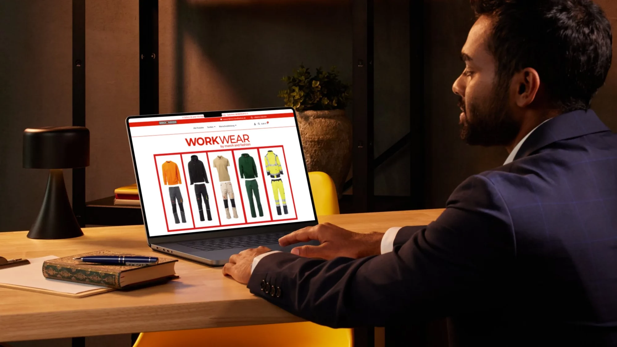 Mann im Anzug am Schreibtisch betrachtet auf dem Laptop den WorkWear-Shop von Merch and Fashion mit Arbeitskleidung; Büro-Szene mit Lampe, Notizbuch und Pflanze.
