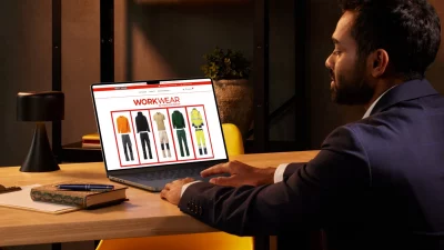 Mann im Anzug am Schreibtisch betrachtet auf dem Laptop den WorkWear-Shop von Merch and Fashion mit Arbeitskleidung; Büro-Szene mit Lampe, Notizbuch und Pflanze.